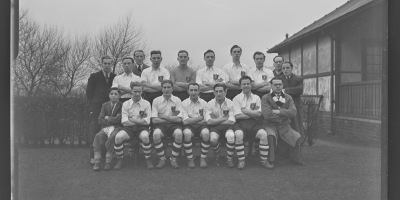 TeampPhotogrpahs of a football team in a white strip with and AA badge and striped socks outside next to a hut